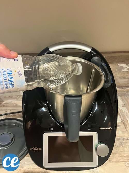 Main tenant une bouteille d'alcool vinaigre cristal près du Thermomix.