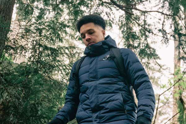 Un homme en doudoune bleue marche pensivement dans une forêt de conifères.