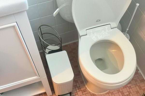 Toilette blanche près d'une poubelle blanche et d'un rangement noir, dans une salle carrelée rose.
