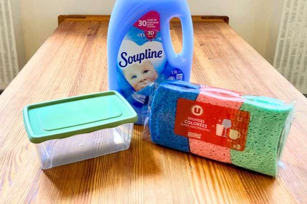 Une boîte Tupperware, de l'adoucissant liquide et des éponges végétales multicolores sur une table en bois.