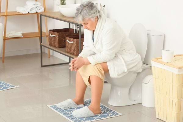Femme âgée assise sur toilettes, regard pensif, en peignoir blanc.