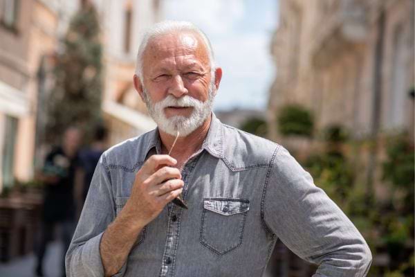 Homme âgé avec une barbe blanche, vêtu d'une chemise grise, pensif dans une rue animée.