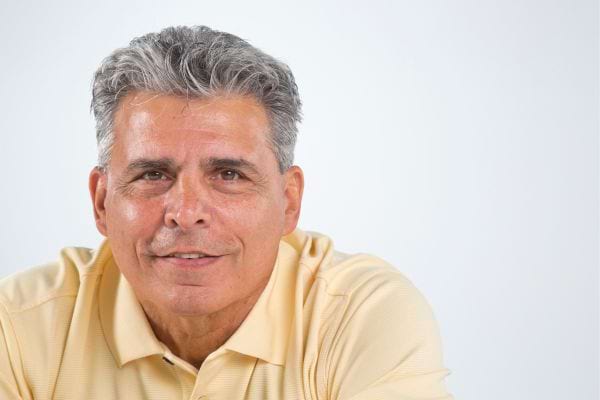 Portrait d'un homme âgé aux cheveux gris portant un polo jaune, souriant à la caméra.