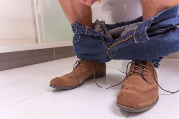 Homme assis sur des toilettes, pantalon baissé, chaussures marron visibles.
