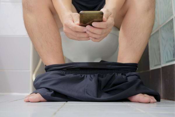 Homme consulte son téléphone, pantalon baissé, sur des toilettes.