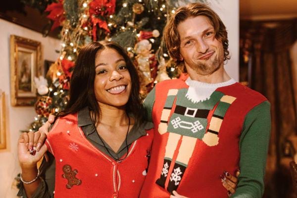 Un couple souriant en pulls de Noël devant un sapin décoré.