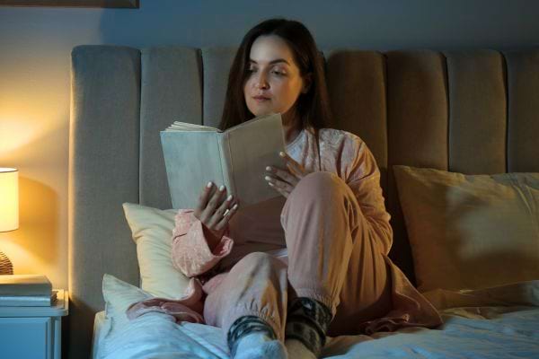 Femme lisant un livre, assise sur un lit éclairé.