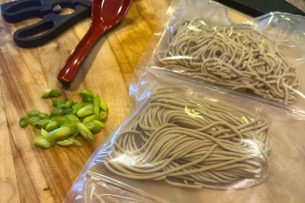 Spaghettis gris dans des sacs sous vide sur une planche à découper avec des oignons verts hachés à côté.