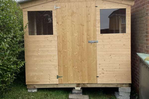Cabanon en bois brut sur parpaings entre maison et haie.