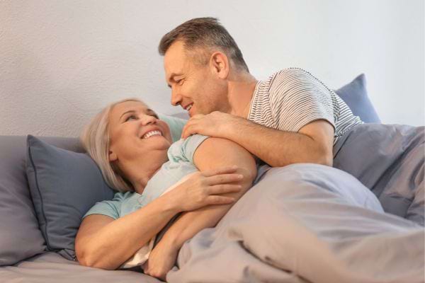 Un couple mature sourit et s’enlace tendrement dans un lit gris.