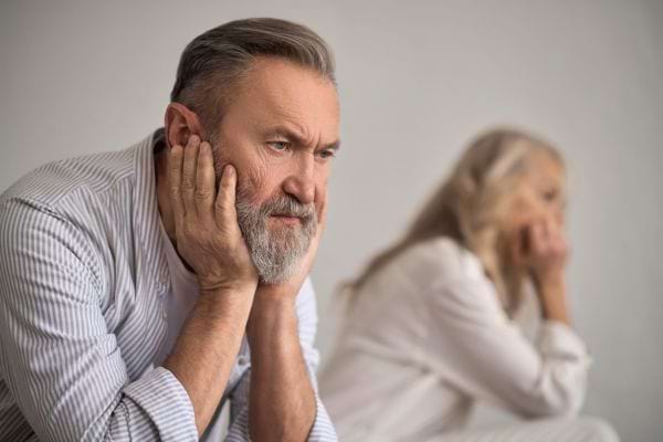 Un homme âgé soucieux, tête entre les mains, une femme en arrière-plan, également pensive.