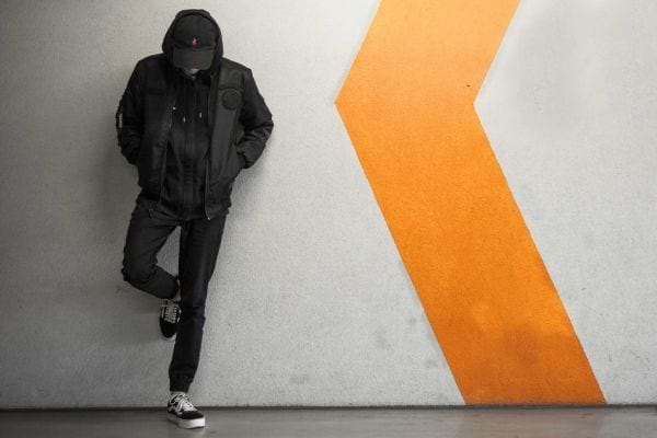 Homme en noir capuche baissée contre un mur avec flèche orange.