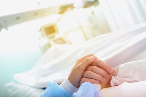 Deux mains se tiennent dans un lit d’hôpital sous une lumière douce.