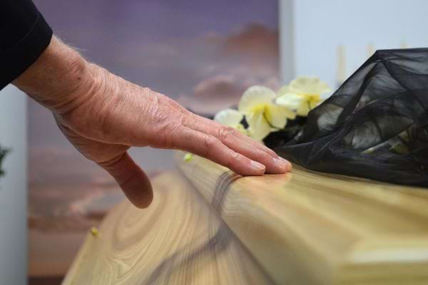 Une main touche doucement un cercueil en bois clair avec des fleurs.