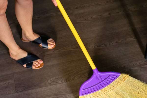 Une personne balaie le sol en bois foncé avec un balai jaune à tête violette, portant des sandales noires.