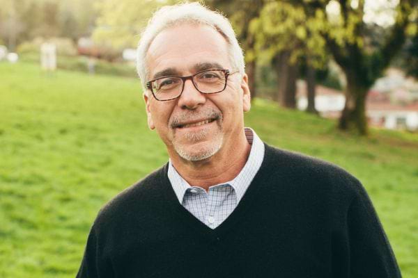 Un homme âgé sourit en extérieur, dans un parc verdoyant.