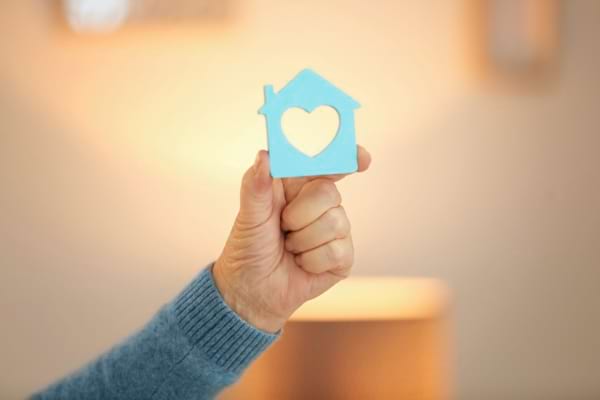 Une main tient une maison bleue avec un cœur découpé.