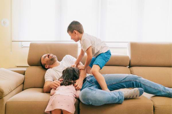 Un père joue joyeusement avec ses deux enfants sur le canapé.