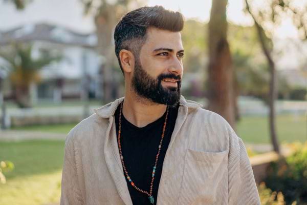 Un homme barbu regarde au loin, debout dans un jardin lumineux.