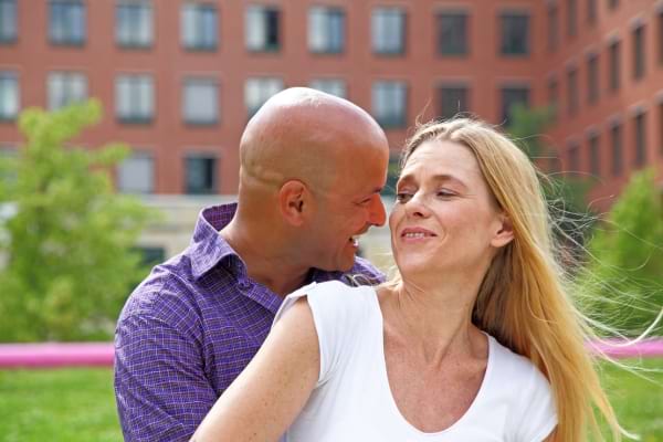 Un couple sourit enlacé dans un parc devant un immeuble.