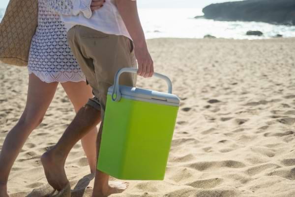 Un couple marche sur la plage, portant une glacière verte pour garder leurs aliments et boissons au frais.