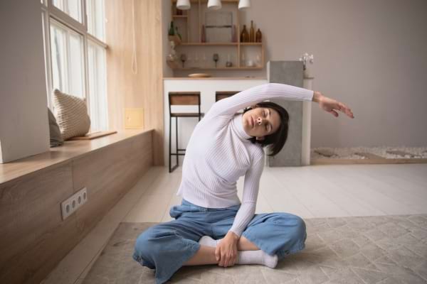 Une femme assise en tailleur fait un étirement latéral dans un salon lumineux.
