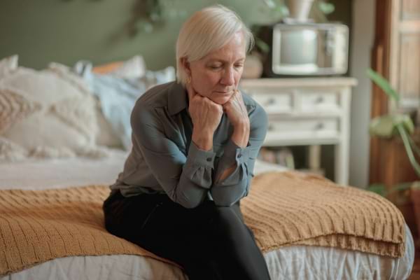 Une femme âgée assise sur un lit semble perdue dans ses pensées.
