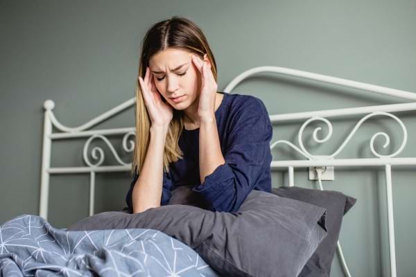 Assise dans son lit, une femme se tient la tête, visiblement gênée par un mal de tête matinal.