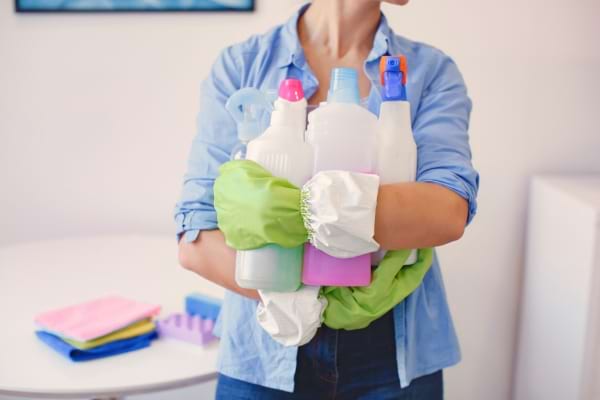 Femme portant des gants et tenant de nombreux produits d’entretien chimiques dans ses bras, prête à faire le ménage.