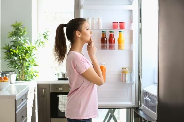 Une femme pensive regarde l’intérieur d’un frigo partiellement rempli de bouteilles.