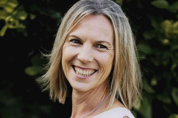 Femme souriante aux cheveux blonds, debout dehors, visage détendu et lumineux.