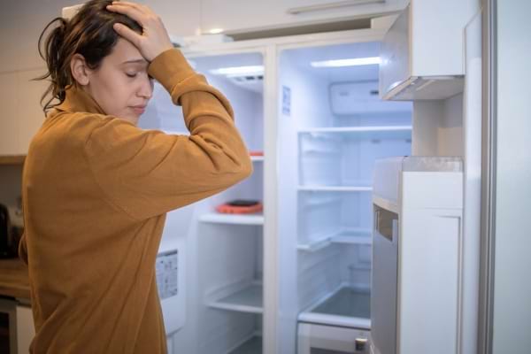 Une femme angoissée découvre son frigo complètement vide, main sur le front.