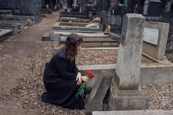 Une femme vêtue de noir s'agenouille devant une tombe, tenant des fleurs rouges.