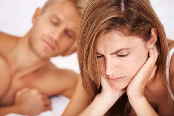 Une femme dans un lit semble triste, l&rsquo;homme derri&egrave;re dort ou r&eacute;fl&eacute;chit.