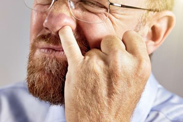 Un homme barbu se cure le nez avec une expression de gêne ou de tension.
