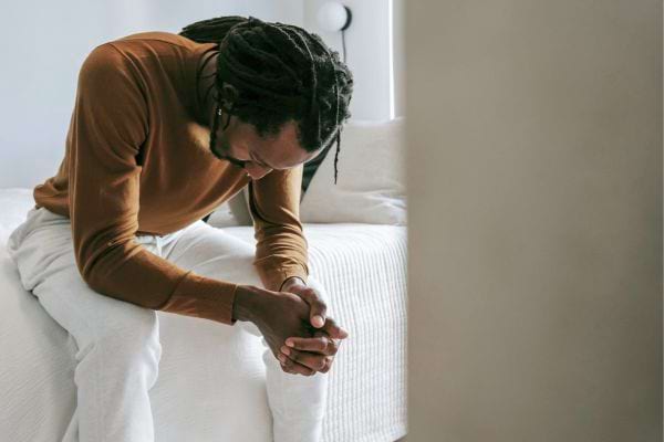 Un homme assis sur un lit, tête baissée, mains jointes.