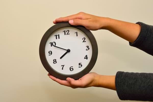 Deux mains tiennent une horloge indiquant environ 9h50.