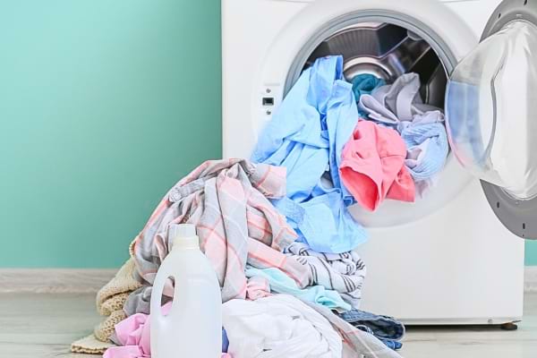 Du linge coloré déborde d’une machine à laver avec une bouteille de lessive posée devant.