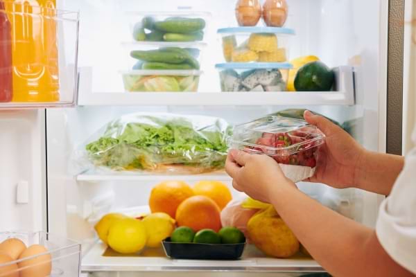 Une personne range un bol de fraises dans un frigo rempli de fruits et légumes frais.