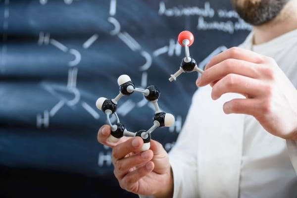 Homme tenant une maquette de molécule devant un tableau noir avec des formules chimiques.