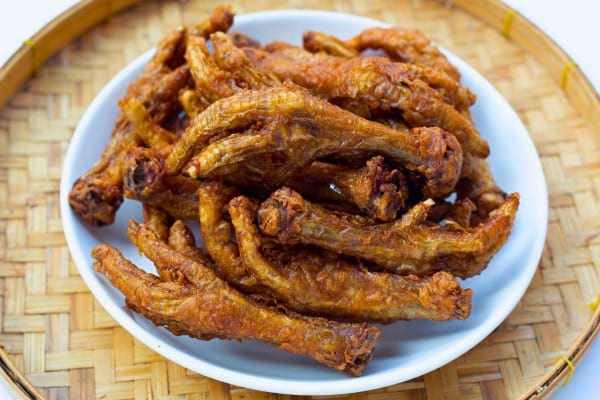 Des pieds de poulet frits sont disposés dans une assiette blanche sur un plateau en bambou.