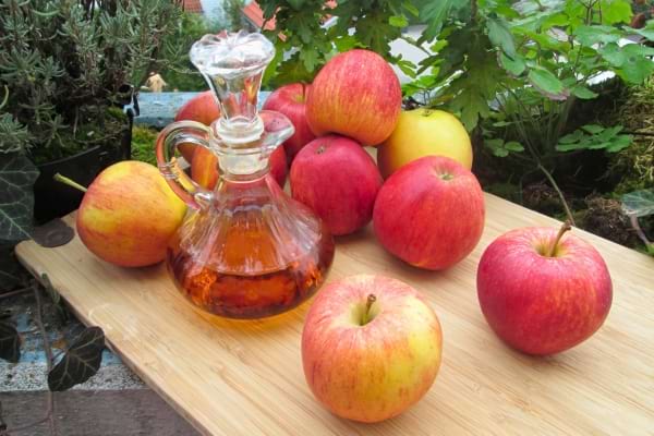 Un flacon de vinaigre de cidre trône au milieu de plusieurs pommes rouges sur une planche.