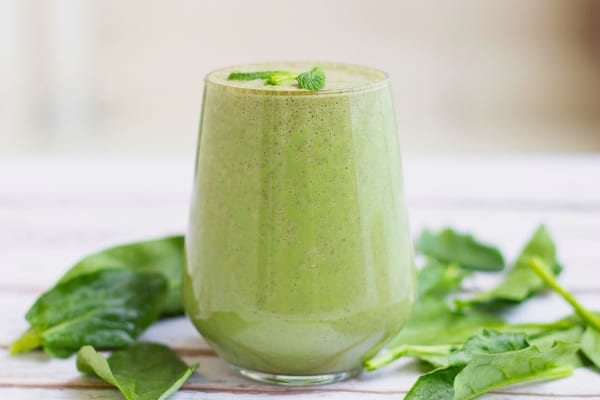 Un smoothie vert dans un verre transparent est entouré de feuilles d’épinard fraîches.