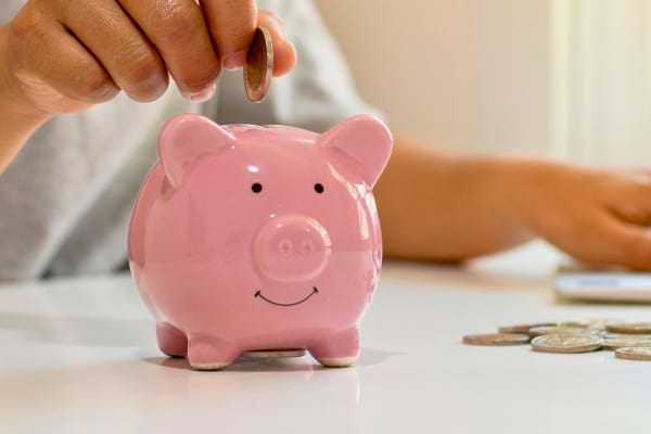 Une main insère une pièce dans une tirelire rose en forme de cochon, illustrant l’idée d’économies.
