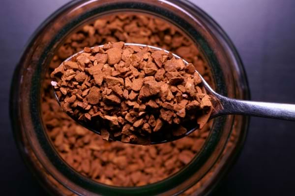 Une cuillère en métal tient du café soluble au-dessus d’un bocal ouvert, rempli de granules foncées.