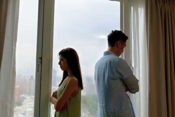 Un couple se tourne le dos devant une fenêtre, chacun les bras croisés, en plein silence tendu.