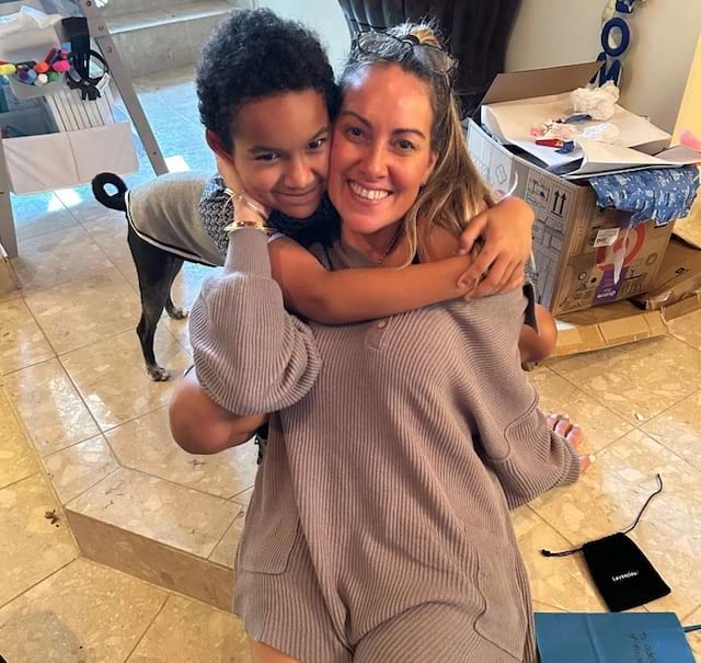 Enfant et femme enlacés avec un chien en arrière-plan.