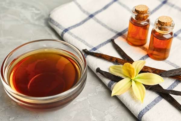 Un bol de liquide ambré repose sur un torchon à carreaux avec des gousses de vanille et une fleur.