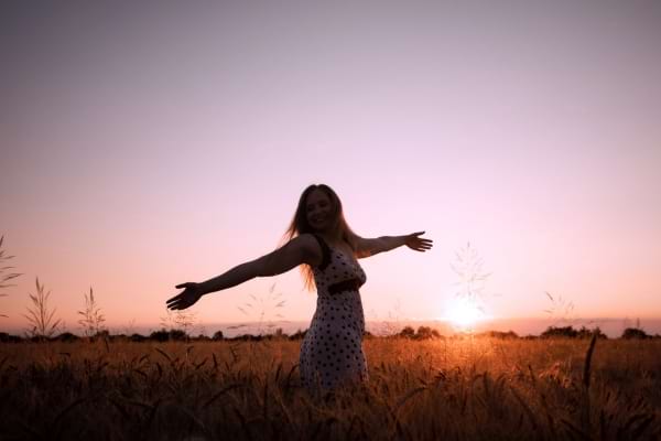 Une femme sourit les bras ouverts dans un champ doré au coucher du soleil.