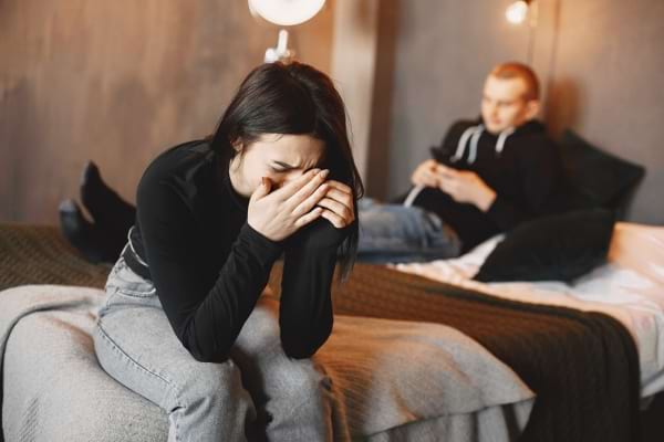 Une femme pleure sur un lit, l’homme derrière elle regarde son téléphone, indifférent.